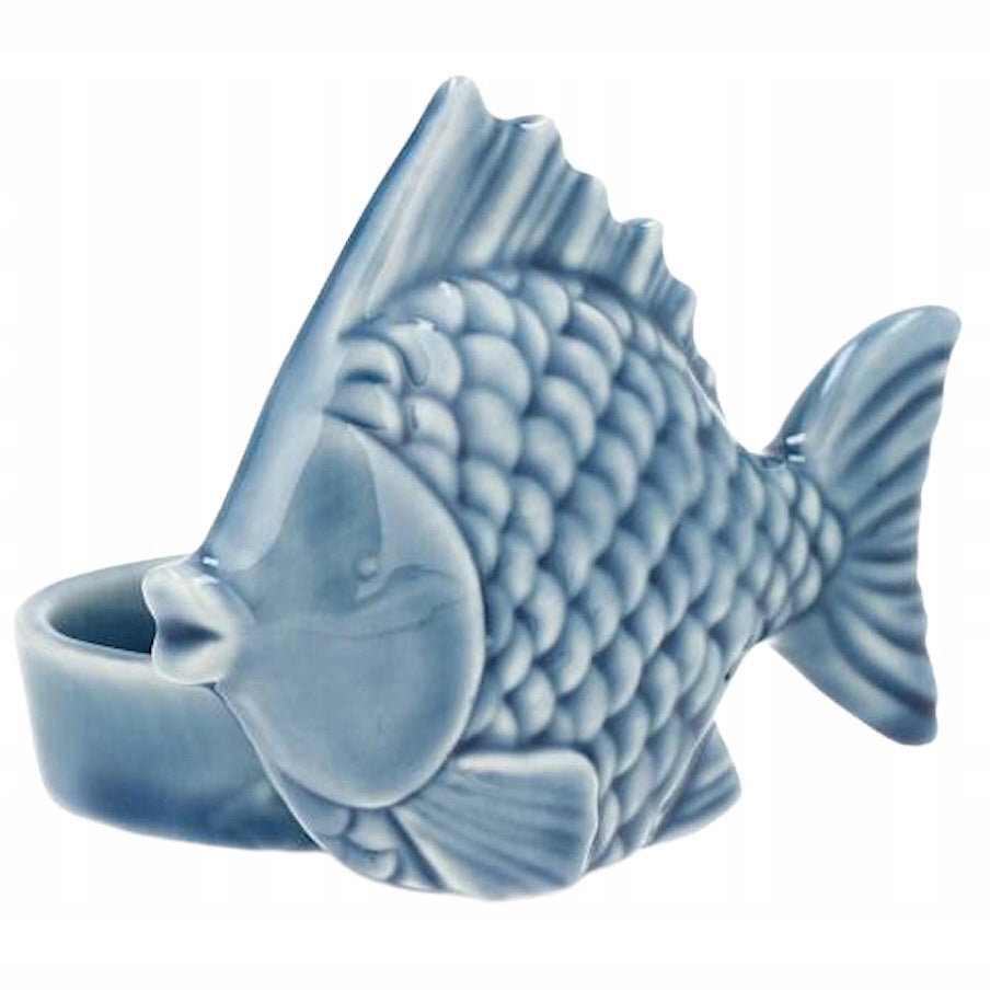 Zdjęcie produktowe na czystym, białym tle (packshot). Niewielki, błękitny świecznik ceramiczny na świeczkę typu tealight. Przednia część świecznika ma formę płaskiej ryby z wyraźnie wytłoczonymi detalami łusek, pyszczka i płetw. Z tyłu za figurką ryby znajduje się okrągły pojemniczek na małą świeczkę. Całość pokryta jest gładkim, błyszczącym szkliwem w pastelowym odcieniu niebieskiego.