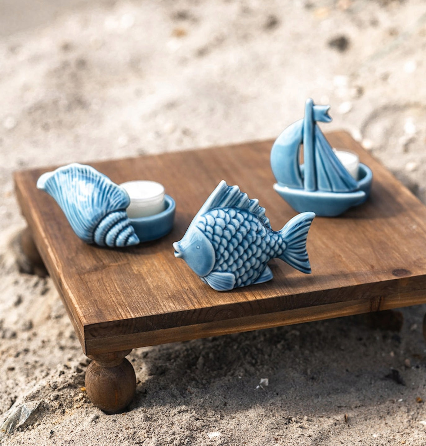 Zbliżenie na trzy małe, błękitne świeczniki ceramiczne na podgrzewacze (tealighty). Są one ułożone na niskim, drewnianym stoliku z toczonymi nóżkami, który stoi bezpośrednio na jasnym piasku. Świeczniki mają urocze, morskie kształty: stożkowatej muszli, pękatej rybki z wyraźnymi łuskami oraz małej żaglówki. Wewnątrz muszli i żaglówki umieszczono białe świeczki.