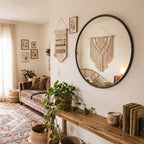 Przytulny salon w stylu boho. Na białej ścianie wisi duże, okrągłe lustro w ciemnej ramie, w którym odbija się wisząca makrama oraz fotel z rattanu. Pod lustrem stoi surowa, drewniana konsola, na której wyeksponowano doniczkę z bujnym epipremnum (pnączem), świecę zapachową i stosik starych książek. Z boku widać brązową, welurową sofę i wzorzysty dywan w stylu vintage.
