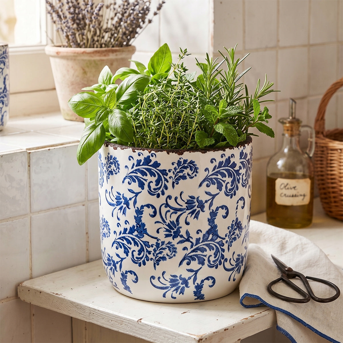 Ceramiczna osłonka w biało-niebieskie wzory z rdzawym, postarzanym rantem, wykorzystana jako pojemnik na świeże zioła (m.in. bazylię, rozmaryn i tymianek). Ustawiona na starym, drewnianym blacie na tle białych, ściennych kafelków. Towarzyszące jej rustykalne nożyczki, butelka z oliwą i pleciony koszyk tworzą sielski, śródziemnomorski klimat tradycyjnej kuchni.