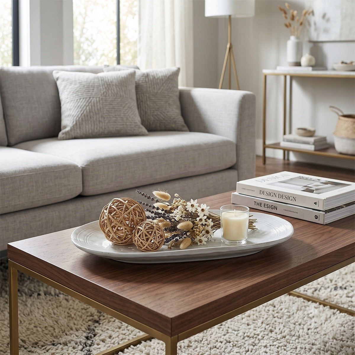 Aranżacja stolika kawowego w eleganckim, nowoczesnym salonie. Na drewnianym blacie stolika leży duża, owalna taca ceramiczna w jasnym kolorze. Ułożono na niej dekoracje: dwie ozdobne kule z rattanu, bukiecik suszonych kwiatów i traw oraz małą, białą świecę w szkle. Obok tacy leżą dwa albumy o designie. W tle widać jasnoszarą kanapę z poduszkami oraz konsolę ze złotymi detalami.