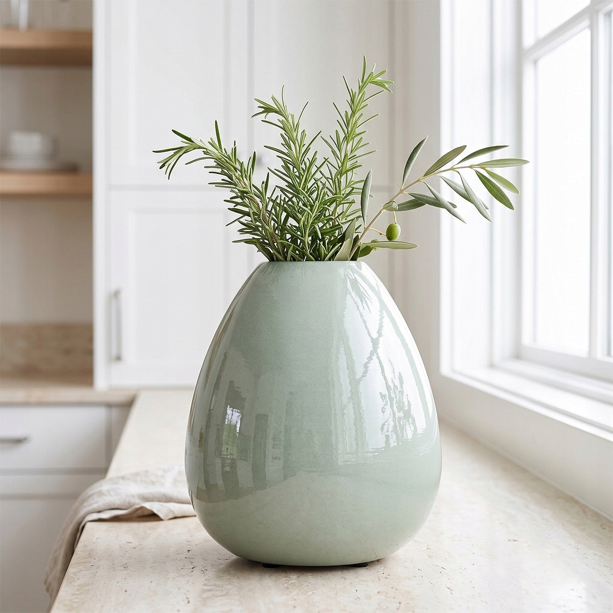 Jasna, elegancka kuchnia z jasnym, kamiennym blatem. Stoi na nim pękaty, jasnozielony wazon ceramiczny o błyszczącej powierzchni, w którym umieszczono świeże gałązki rozmarynu oraz oliwki. W tle widać okno, białe szafki kuchenne oraz rozmyte drewniane półki, co nadaje całości świeży, śródziemnomorski charakter.