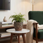 Przytulny kącik wypoczynkowy w salonie w stylu mid-century modern. Na małym stoliku z jasnym, marmurowym blatem i drewnianymi nóżkami stoi patynowana doniczka z wytłaczanym wzorem, obsadzona zieloną paprocią. Obok znajduje się fotel obity ciemnozielonym welurem, na którym swobodnie leży jasny, dziergany koc. Pod blatem stolika widać stos eleganckich albumów i książek.