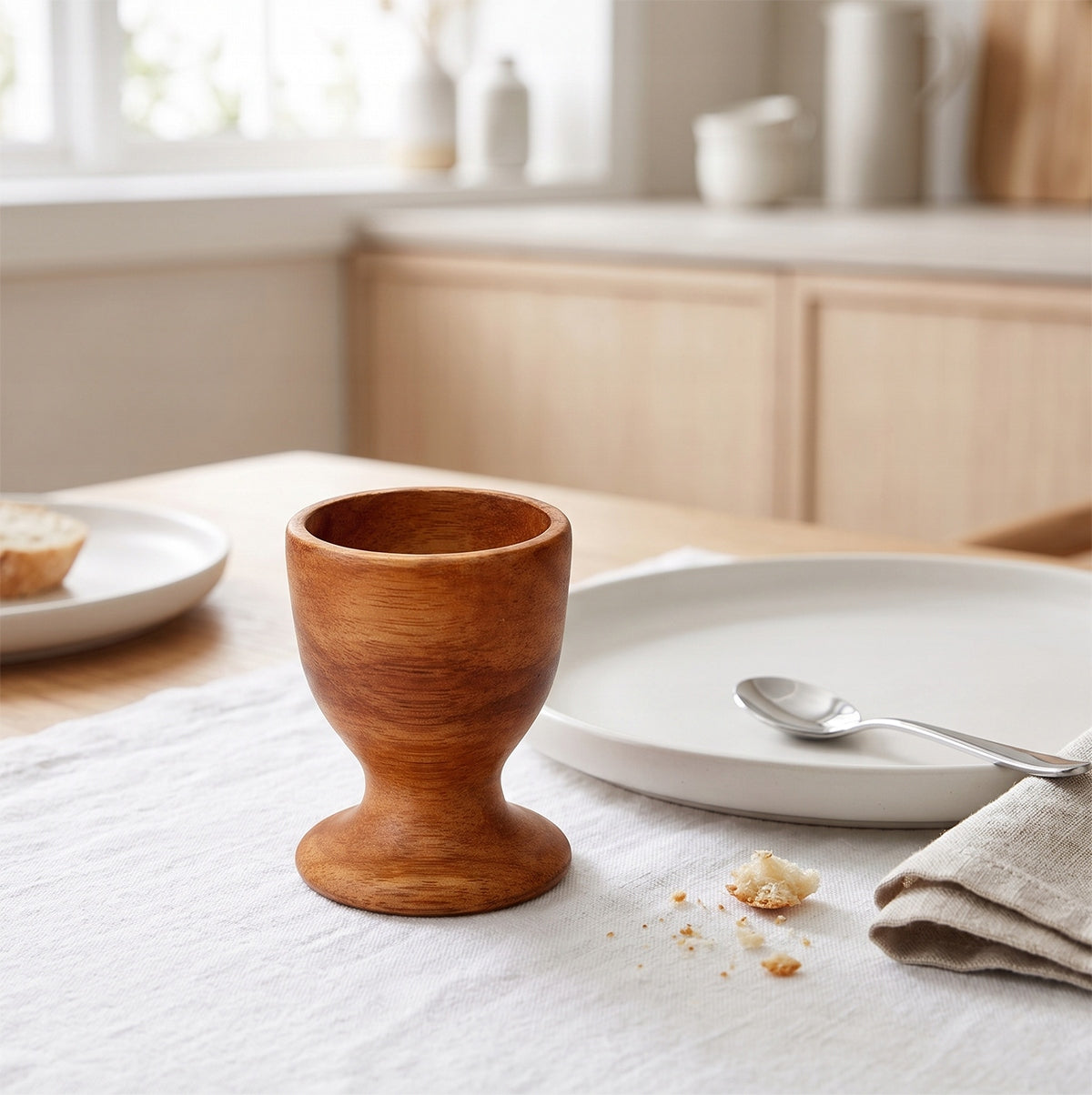 Pusty, drewniany kieliszek na jajko ustawiony na stole przykrytym białym, lnianym obrusem. Obok podstawki zaaranżowano minimalistyczne nakrycie: jasny, ceramiczny talerz z ułożoną obok małą łyżeczką oraz lnianą serwetkę. W rozmytym tle widać nowoczesne, jasne meble kuchenne, co podkreśla skandynawski charakter stylizacji.