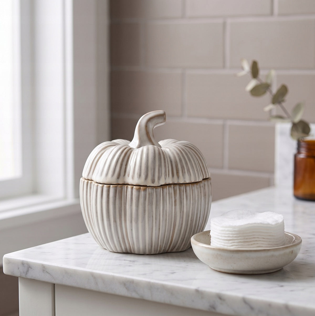 Jasna aranżacja łazienkowa w naturalnym stylu. Na marmurowym blacie wyeksponowano kremowy, prążkowany pojemnik ceramiczny w kształcie dyni z przykrywką. Tuż obok, na małym ceramicznym spodeczku, leży stosik białych wacików kosmetycznych. W tle widać jasne kafelki, okno oraz ciemny, szklany słoiczek z gałązką eukaliptusa.