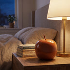 Nastrojowa sypialnia uchwycona o zmroku. Na drewnianej szafce nocnej z wyraźnym rysunkiem słojów stoi dekoracyjne, ceramiczne jabłko w pomarańczowym kolorze. Obok ułożony jest stosik czterech książek. Scenę oświetla miękkie, nastrojowe światło z eleganckiej lampy nocnej ze złotą podstawą i jasnym abażurem. W tle widoczne jest zaścielone łóżko z szarą, lnianą pościelą oraz okno, za którym widać granatowe, wieczorne niebo.