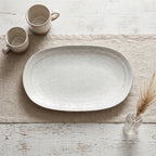Ujęcie z góry (flatlay) na blat ze starego, bielonego drewna z wyraźnymi przetarciami. W centrum, na beżowym lnianym bieżniku, leży pusty, owalny talerz ceramiczny o jasnoszkliwionej, organicznej powierzchni. Kompozycję uzupełniają dwa małe, dwukolorowe kubki ceramiczne w lewym górnym rogu oraz malutki, szklany wazonik z suszoną trawą w prawym dolnym rogu.
