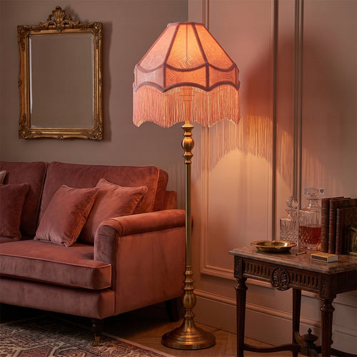 Podświetlony, jasnoróżowy abażur z frędzlami w stylu retro, zamontowany na wysokiej, mosiężnej lampie podłogowej. Lampa stoi w nastrojowym salonie obok różowej, welurowej sofy. W tle widoczne jest ozdobne lustro w złotej ramie oraz stylowy stolik z karafkami i książkami. Ciepłe światło lampy tworzy przytulną atmosferę we wnętrzu