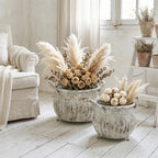 Jasne, romantyczne wnętrze w stylu shabby chic. Dwie duże, przecierane donice z metalu na małych nóżkach stoją na białej, drewnianej podłodze. Są bogato wypełnione kompozycją z suszonych kwiatów: beżowych róż, liści eukaliptusa oraz puszystej trawy pampasowej. W tle po lewej stronie stoi jasny fotel z plecionym kocem, a po prawej drewniana drabinka z mniejszymi doniczkami.