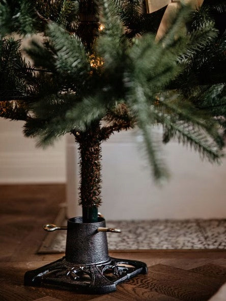 Zbliżenie na dolną część choinki osadzonej w ozdobnym, czarnym stojaku żeliwnym. Widoczny jest pień drzewka oraz masywna podstawa stojaka z ażurowym wzorem i wkręconymi śrubami (uszami) mocującymi. Ozdoba stoi na eleganckim, drewnianym parkiecie ułożonym w jodełkę.