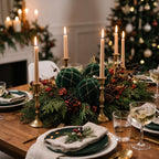 Eleganckie nakrycie stołu wigilijnego w klasycznym stylu. Środek drewnianego blatu zdobi bogaty stroik z naturalnych gałązek iglastych, szyszek i suszonych owoców, w którym wyeksponowano trzy ciemnozielone, welurowe bombki ze złotą kratką. W kompozycję wpleciono mosiężne świeczniki ze smukłymi, zapalonymi świecami, a po bokach widać kryształowe kieliszki oraz talerze z zielonymi serwetkami. W rozmytym tle błyszczą lampki na ubranej choince.