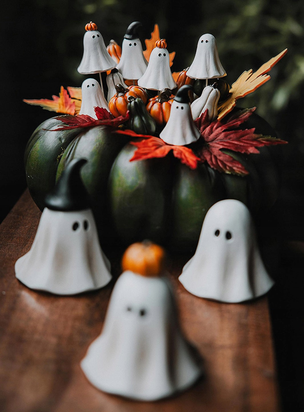 Nastrojowa, mroczna kompozycja na Halloween. Na ciemnym, drewnianym blacie stoi duża, ciemnozielona dynia pełniąca funkcję bazy stroika. Wyrastają z niej sztuczne, jesienne liście w intensywnych odcieniach czerwieni i żółci, małe dynie oraz liczne piki dekoracyjne w kształcie białych duszków (część z nich ma czarne kapelusze lub dynie na głowach). Na pierwszym, mocno rozmytym planie stoją bezpośrednio na blacie trzy dodatkowe figurki duszków.