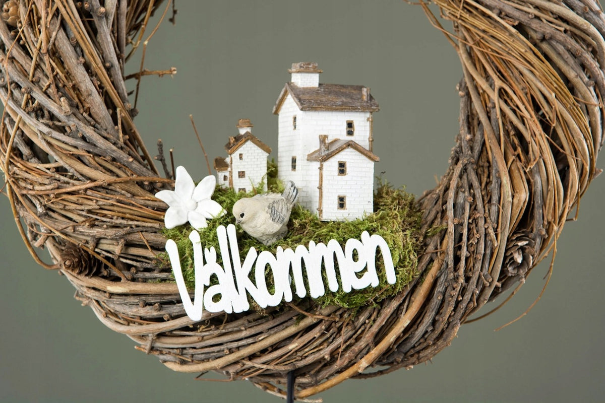 Zbliżenie na rustykalny wieniec spleciony z grubych, naturalnych gałęzi. Wewnątrz wieńca znajduje się kompozycja z mchu, na którym ustawiono figurkę białej, piętrowej kamieniczki z małymi okienkami, figurkę małego ptaszka oraz ceramiczny kwiatek. U dołu kompozycji wkomponowano biały, wycięty napis "Välkommen".
