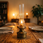Zapalona mosiężna lampa naftowa z wysokim, ryflowanym kloszem ze szkła, ustawiona na środku dużego, drewnianego stołu jadalnianego. Płomień rzuca na blat wyraziste, gwiaździste cienie. Po bokach widoczne są rozmyte nakrycia stołowe z talerzami i serwetkami, a w tle majaczą inne, ciepłe źródła światła, budując przytulną atmosferę wieczoru.