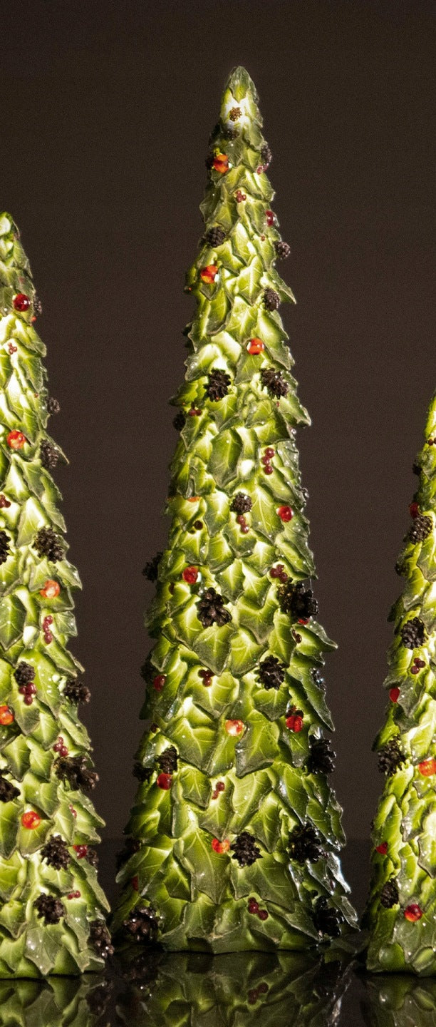 Efektowna, wieczorna kompozycja prezentująca trzy dekoracyjne choinki z motywem liści. Na tym ujęciu figurki są wewnętrznie podświetlone, co w piękny sposób wydobywa głęboką zieleń, podkreśla krawędzie liści i sprawia, że czerwone koraliki lśnią niczym drobne lampki. Ozdoby ustawione są na ciemnym, błyszczącym blacie, który delikatnie odbija ich ciepły blask, tworząc magiczny, przytulny klimat idealny na zimowe wieczory.