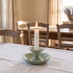 Przytulna jadalnia w wiejskim stylu. Na stole nakrytym jasnym, lnianym obrusem stoi ceramiczny świecznik w kolorze chłodnego, jasnego błękitu/zieleni (celadon). Świecznik ma formę miseczki z umieszczoną pośrodku wyższą podstawką, w której płonie wysoka, biała świeca. W rozmytym tle widać drewniane krzesła i stół oraz jasne zasłony w oknie.