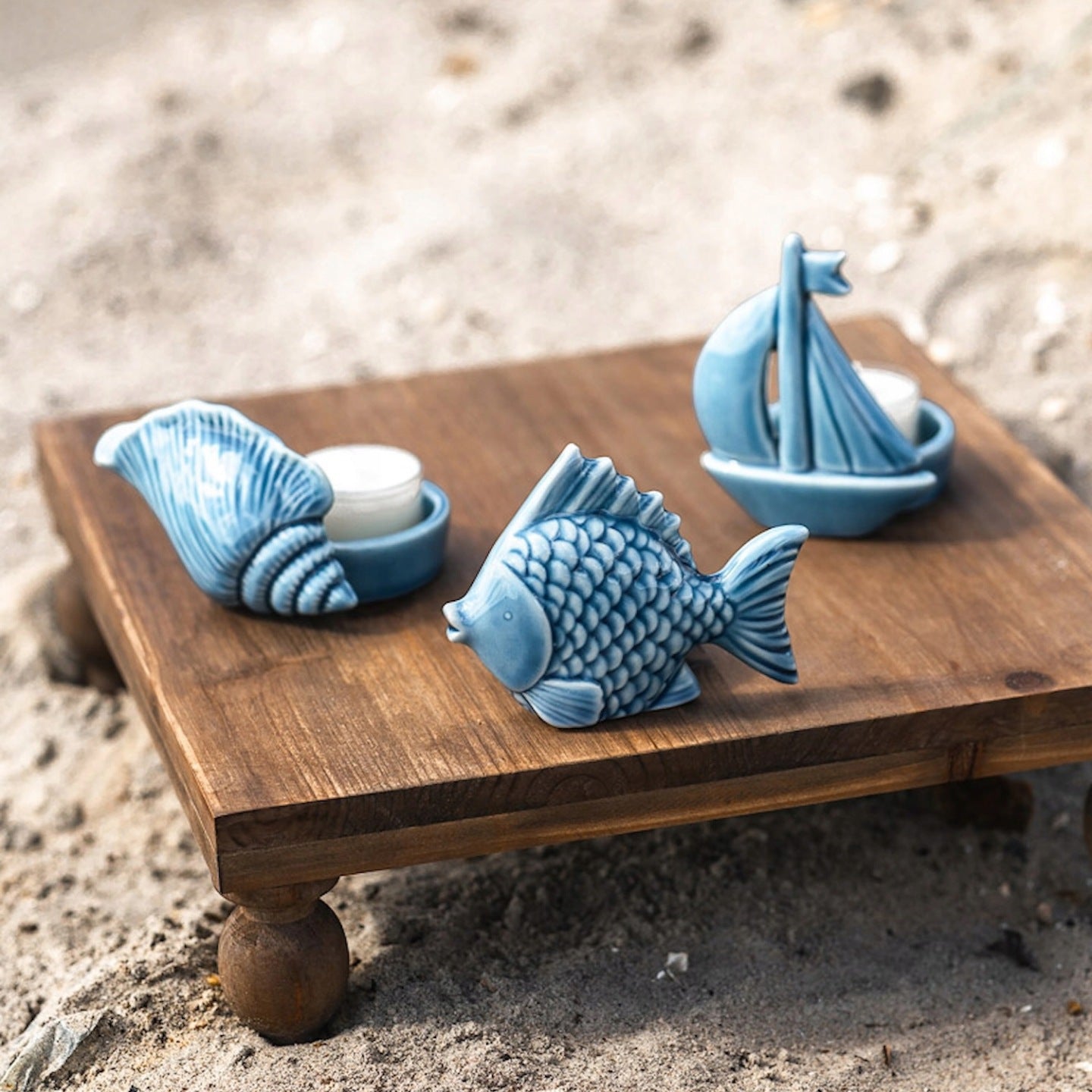 Zestaw niebieskich, ceramicznych dekoracji marynistycznych ustawionych na małym, kwadratowym drewnianym stoliku z toczonymi nóżkami, stojącym na plaży. Wśród nich znajduje się figurka ryby oraz dwa świeczniki na tealighty: jeden w kształcie żaglówki, drugi przypominający dużą muszlę z wydzielonym miejscem na świecę.