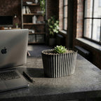 Nowoczesne biuro domowe (home office) w stylu loft. Na betonowym blacie biurka, tuż obok otwartego laptopa i pióra, stoi owalna, żłobiona doniczka w ciemnoszarym kolorze. Wypełniona jest drobnym żwirkiem, a w centrum rośnie mały, zielony sukulent. W głębi widać industrialne okna i ceglane ściany.