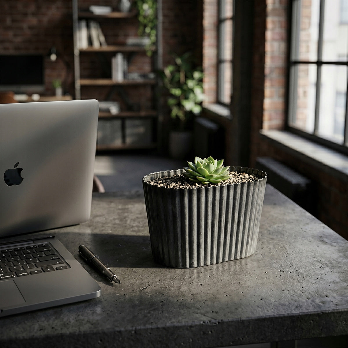 Nowoczesne biuro domowe (home office) w stylu loft. Na betonowym blacie biurka, tuż obok otwartego laptopa i pióra, stoi owalna, żłobiona doniczka w ciemnoszarym kolorze. Wypełniona jest drobnym żwirkiem, a w centrum rośnie mały, zielony sukulent. W głębi widać industrialne okna i ceglane ściany.