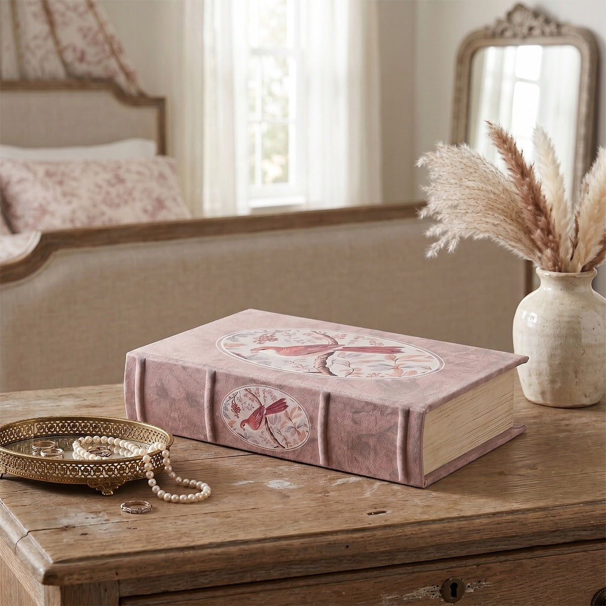 Romantyczna aranżacja sypialni w stylu shabby chic. Na starej, drewnianej komodzie z widocznym usłojeniem i dziurką od klucza leży różowe pudełko-książka z florystycznym motywem ptaka. Po lewej stronie stoi złota, ażurowa tacka z dnem z lustra, na której leży elegancki sznur pereł i pierścionki. Z prawej strony znajduje się jasny, postarzany wazonik z suszonymi, puszystymi trawami. W jasnym, rozmytym tle widać łóżko z pościelą w drobne kwiaty i lustro powieszone na ścianie.