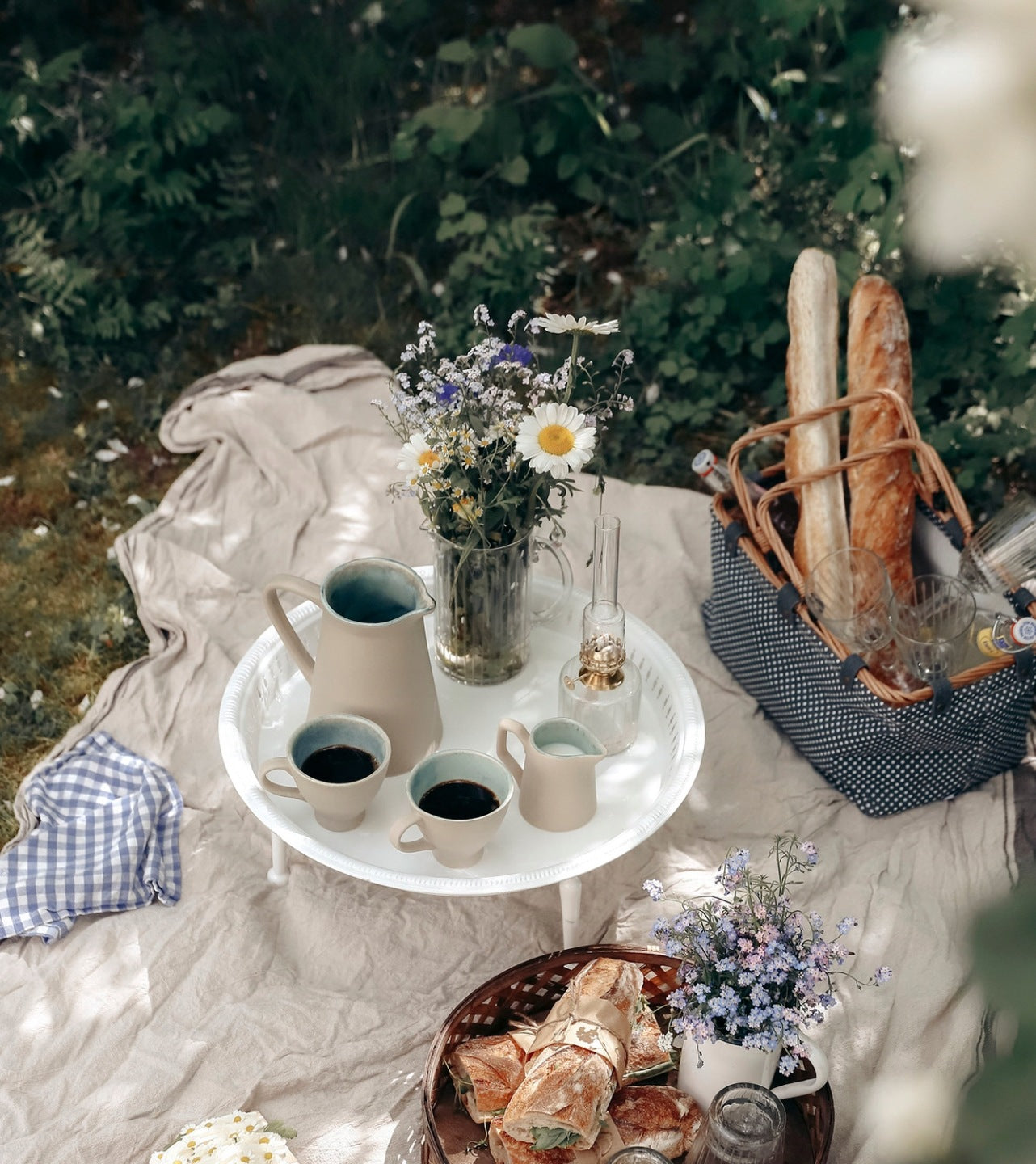 Estetyczny piknik na jasnym kocu rozłożonym w cieniu na trawie. W centrum znajduje się okrągły, biały stolik-taca, na którym stoi beżowy ceramiczny dzbanek, mlecznik, dwie filiżanki z czarną kawą, szklany wazon z polnymi kwiatami (rumianki, chabry) oraz mała, niezalona lampa naftowa. Dookoła zaaranżowano kosz piknikowy z bagietkami i kieliszkami, a na pierwszym planie tackę z kanapkami. Sceneria utrzymana w sielskim, letnim klimacie
