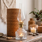 Przytulne wnętrze w stylu boho. Na drewnianej komodzie stoi zapalony szklany świecznik w kształcie lampy naftowej z prążkowanego szkła. Wokół niego zaaranżowano naturalne dekoracje: plecione, rattanowe pojemniki i wazony, gładkie kamienie rzeczne oraz prostą świecę w szkle. W tle na jasnej ścianie wisi beżowa makrama, a po prawej stronie widać fragment rośliny doniczkowej.