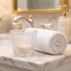 Jasna, elegancka łazienka zaaranżowana w stylu domowego spa. Na jasnym, marmurowym blacie umywalki stoi szklany świecznik z tłoczonym wzorem w romby. Wewnątrz znajduje się mała, niezapalona świeczka w jasnym kolorze. Tuż obok świecznika leży starannie zwinięty w rulon, gruby, biały ręcznik frotte. W rozmytym tle widać baterię umywalkową oraz dozownik na mydło z elementami w kolorze złota/mosiądzu.