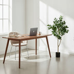 Minimalistyczny, bardzo jasny kącik do pracy (home office). W rogu pokoju, obok prostego, drewnianego biurka w stylu skandynawskim, stoi sztuczne drzewko figowe w czarnej donicy. Na biurku znajduje się szary laptop, książki, notes oraz ceramiczny kubek. Do biurka wsunięte jest jasne, tapicerowane krzesło na metalowych nogach. Światło z dużego okna rzuca wyraźne cienie na białe ściany.