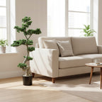 Przestronny, nowoczesny salon z dużymi oknami. W rogu, obok eleganckiej, jasnobeżowej sofy, stoi imponujące, sztuczne drzewko bonsai, imitujące sosnę. Roślina ma fantazyjnie wygięty, gruby pień i charakterystyczne, ułożone w chmurki igły. Jest posadzona w czarnej, prostej donicy. Wnętrze jest jasne, oświetlone naturalnym, wpadającym przez okna słońcem, które rzuca delikatne cienie.
