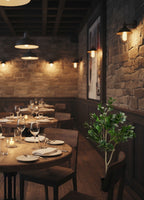 Nastrojowe wnętrze eleganckiej restauracji w stylu industrialnym/loftowym. Pomiędzy drewnianymi, okrągłymi stołami z przygotowanymi nakryciami i płonącymi świecami stoi sztuczne drzewko figowe z ciemnozielonymi liśćmi, ożywiając przestrzeń. Wnętrze charakteryzuje się ścianą z surowego, ciosanego kamienia lub cegły, na której zawieszono stylowe, metalowe kinkiety dające ciepłe, przytłumione światło.
