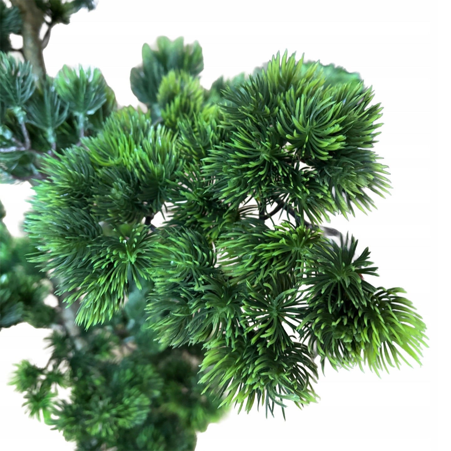 Makrofotografia (zbliżenie) na gałązki sztucznego drzewka bonsai. Obraz ostro i szczegółowo ukazuje strukturę intensywnie zielonych, plastikowych igieł, zebranych w gęste pęczki (chmurki) naśladujące naturalny wygląd sosny. Tło jest całkowicie białe, co podkreśla wyrazistość igliwia.