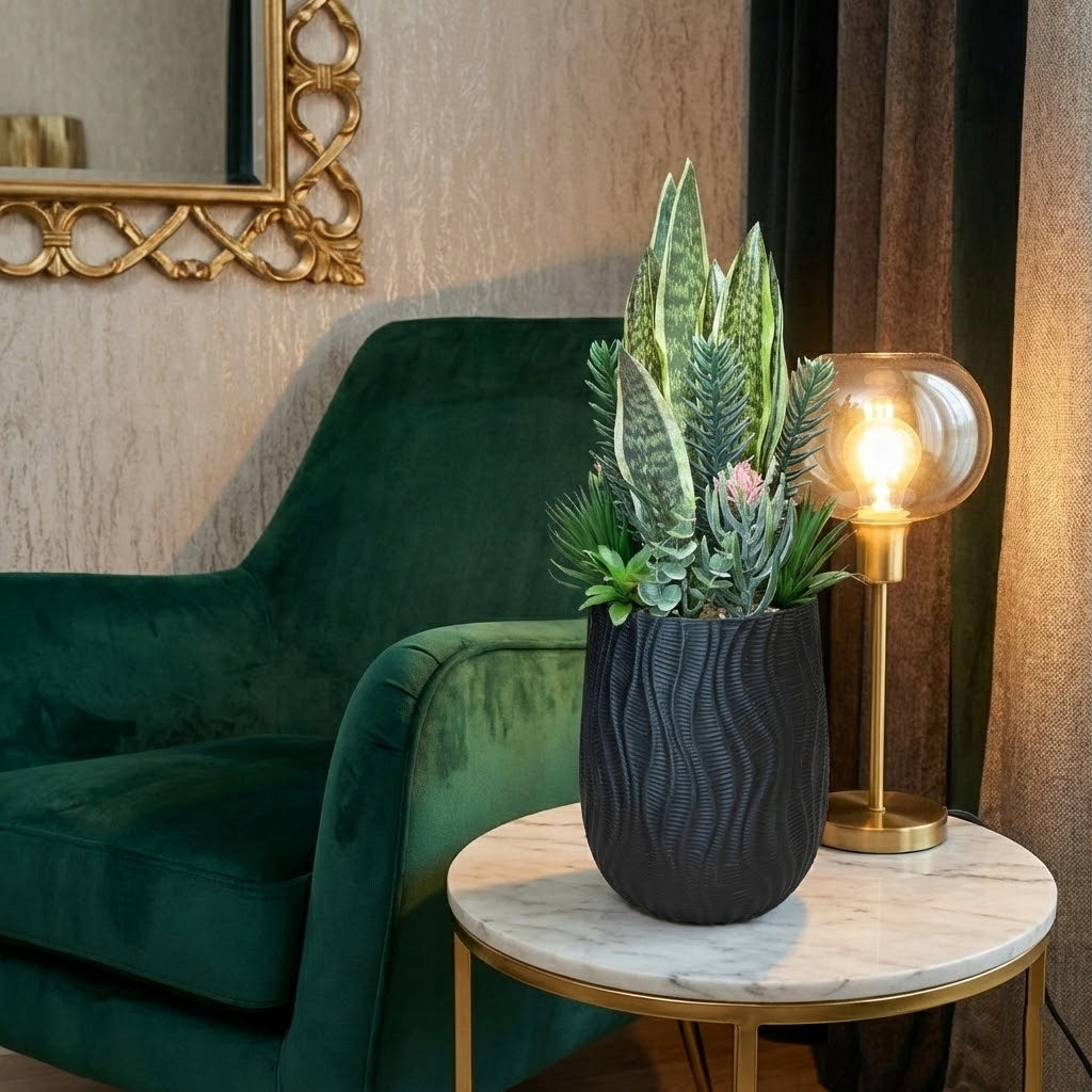 Elegancki, nastrojowy salon w stylu glamour. Na małym, marmurowym stoliku ze złotymi nogami umieszczono kompozycję sztucznych sukulentów i sansewierii w czarnej doniczce we wzór fali. Obok rośliny stoi zapalona lampka nocna ze szklanym, bursztynowym kloszem. W tle znajduje się ciemnozielony, welurowy fotel oraz ściana z ozdobnym lustrem w złotej ramie