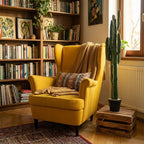 Przytulny salon w stylu vintage z dużą, drewnianą biblioteczką pełną książek. W centrum kadru stoi musztardowo-żółty fotel uszak z zarzuconym kocem i wzorzystą poduszką. Tuż obok, na odwróconej drewnianej skrzynce, wyeksponowano wysokiego, sztucznego kaktusa w czarnej doniczce. Wnętrze oświetla ciepłe, naturalne światło wpadające przez okno po prawej stronie.