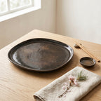 Ciemny, rustykalny talerz ceramiczny w zupełnie nowej, jasnej odsłonie w minimalistycznym stylu Japandi. Naczynie ustawiono na gładkim, dębowym stole w towarzystwie bambusowych pałeczek opartych na gładkim kamyku oraz małej miseczki na sos. Subtelności dodaje jasna, lniana serwetka z delikatną gałązką kwitnącej wiśni. Zestawienie ciemnego, lśniącego szkliwa z jasnym drewnem i miękkim światłem dziennym daje bardzo świeży, naturalny efekt.