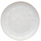 Zdjęcie produktowe typu packshot wykonane z góry (flat lay) na czystym, białym tle. Przedstawia pusty, okrągły talerz ceramiczny o płytkim profilu. Naczynie charakteryzuje się jasną, kremowo-szarą kolorystyką z bardzo delikatnym, nakrapianym deseniem (przypominającym szkliwo reaktywne) oraz subtelnie zaznaczonym, beżowym rantem. Minimalistyczny i naturalny wygląd sprawia, że jest to idealna baza do serwowania różnorodnych potraw.