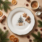 Zdjęcie z góry (flat lay) na drewnianym stole w świątecznym klimacie. W centrum znajduje się biały, ceramiczny talerz z falowanym brzegiem i zieloną obwódką, ozdobiony w środku ilustracją ubranej choinki w koszu z prezentami. Na talerzu leży pierniczek w kształcie gwiazdki oraz orzechy włoskie. Wokół zaaranżowano gałązki świerku, laski cynamonu, anyż gwiaździsty, plastry suszonej pomarańczy oraz kubek kawy z cynamonem i mały dzbanuszek na mleko.