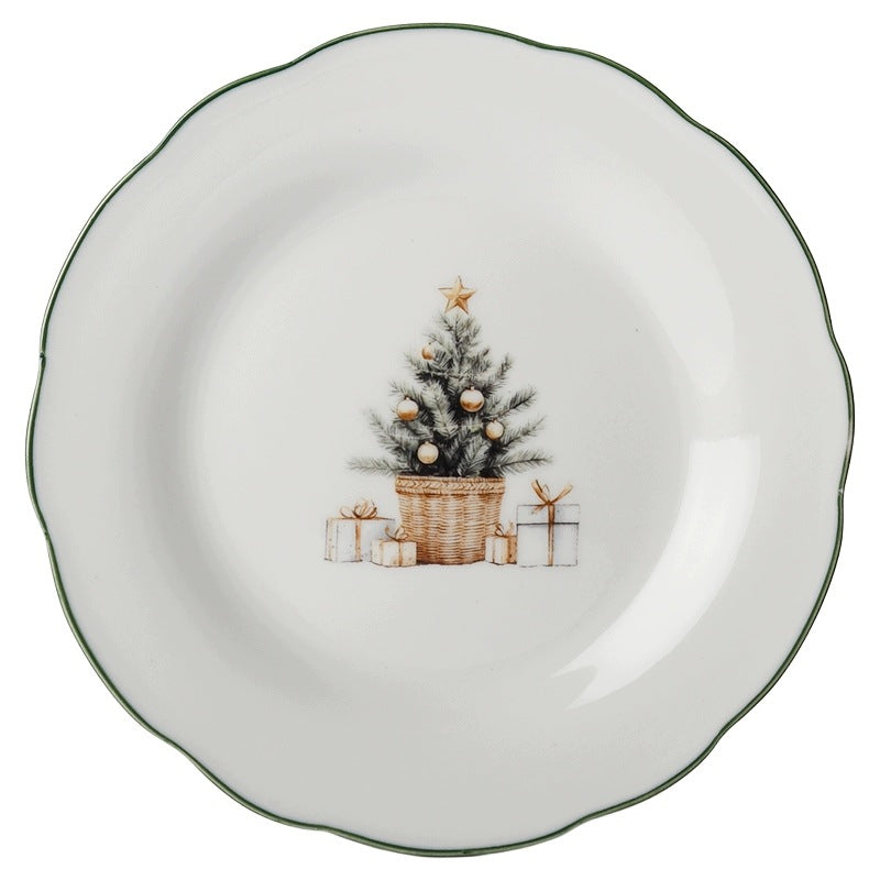 Zdjęcie produktowe na gładkim, białym tle (packshot). Prezentuje okrągły, ceramiczny talerz deserowy o pofalowanym brzegu podkreślonym wąską, ciemnozieloną linią. W centralnej części umieszczona jest subtelna grafika przedstawiająca ubraną choinkę osadzoną w plecionym koszu, ze złotą gwiazdą na wierzchołku, u której podstawy leżą starannie przewiązane prezenty.