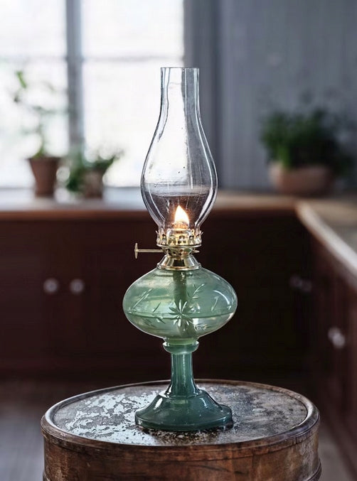 Zapalona, tradycyjna lampa naftowa w stylu vintage, stojąca na starym, drewnianym taborecie z łuszczącą się farbą. Podstawa i zbiornik na naftę wykonane są z grubego, matowego szkła w kolorze morskiej zieleni, ozdobionego rzeźbionym wzorem gwiazd i gałązek. Mechanizm palnika jest mosiężny, z widocznym pokrętłem do regulacji knota. Na górze znajduje się przezroczysty, smukły, szklany komin. W rozmytym tle widać rustykalną kuchnię z drewnianymi szafkami i oknem, przez które wpada naturalne światło.