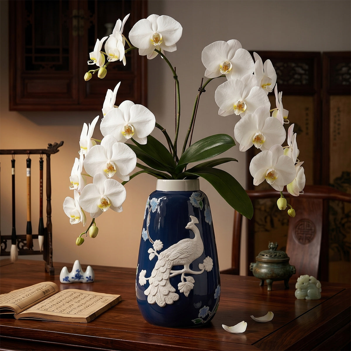 Granatowy wazon z białym reliefem pawia, w którym umieszczono imponującą gałązkę białego storczyka (orchidei). Naczynie stoi na masywnym, ciemnodrewnianym biurku w otoczeniu orientalnych dodatków: stojaka z pędzlami do kaligrafii, otwartej księgi z chińskimi znakami, małego kadzielnika oraz ceramicznych figurek. Tło z drewnianym parawanem podkreśla elegancki, azjatycki styl aranżacji.