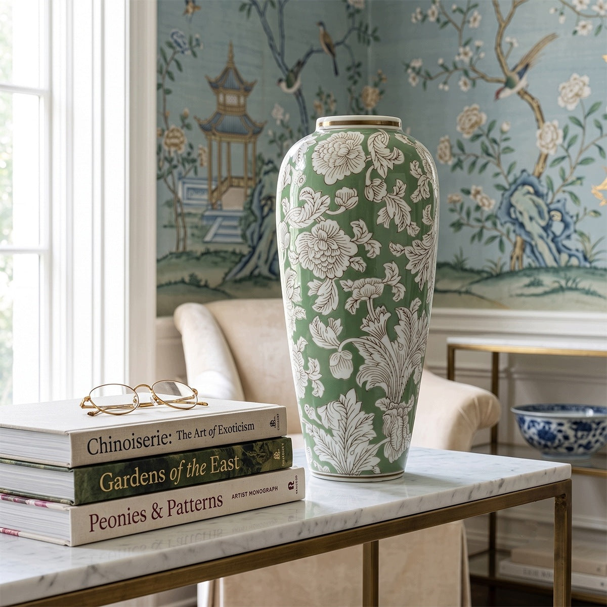 Jasny kącik w salonie w stylu chinoiserie. Na stoliku z białym, marmurowym blatem i mosiężną ramą stoi wysoki, zielony wazon ceramiczny z białym wzorem kwiatowym. Obok wazonu leży stos trzech książek o tematyce sztuki i ogrodów (m.in. "Chinoiserie: The Art of Exoticism"), na których spoczywają złote okulary. W tle znajduje się fotel oraz tapeta ścienna przedstawiająca chiński pejzaż z pagodą, kwitnącymi drzewami i ptakami.