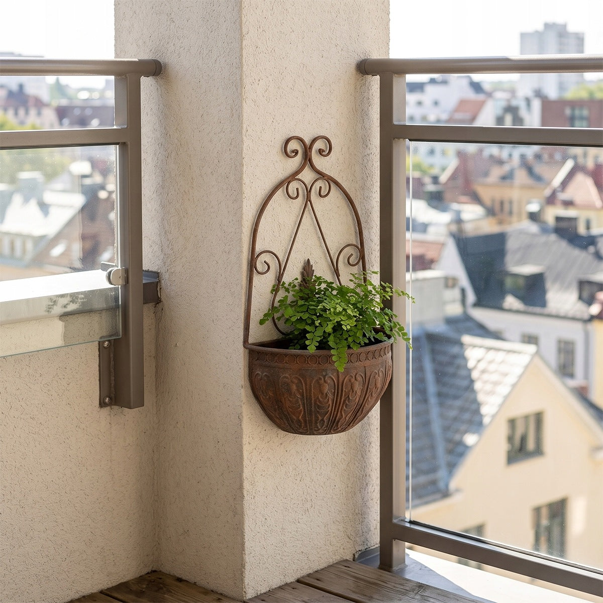 Aranżacja na jasnym, słonecznym balkonie z nowoczesną, przeszkloną balustradą z metalowymi poręczami. Na białym, tynkowanym filarze zawieszono dekoracyjną, półokrągłą donicę ścienną z metalu o rdzawym, postarzanym wykończeniu i kutych zdobieniach. Wewnątrz naczynia rośnie świeża, jasnozielona paproć. W tle za barierką rozpościera się widok na miejską zabudowę z pastelowymi elewacjami i spadzistymi dachami.