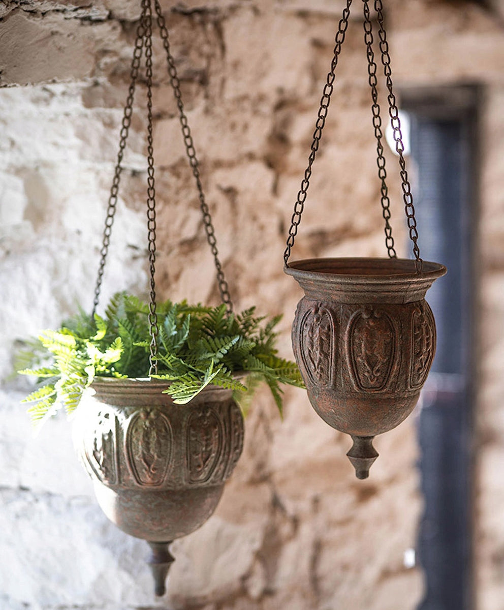 Rustykalna aranżacja wnętrza lub zadaszonego tarasu. Na tle surowej, jasnej ściany z ciosanego kamienia wiszą dwie metalowe donice w stylu vintage, pokryte rdzawą patyną, zawieszone na cienkich łańcuszkach. Lewa donica wypełniona jest soczyście zieloną paprocią, natomiast prawa donica jest pusta, co pozwala dokładnie zobaczyć jej owalny, tłoczony wzór i dekoracyjne, dolne zwieńczenie w kształcie łezki.