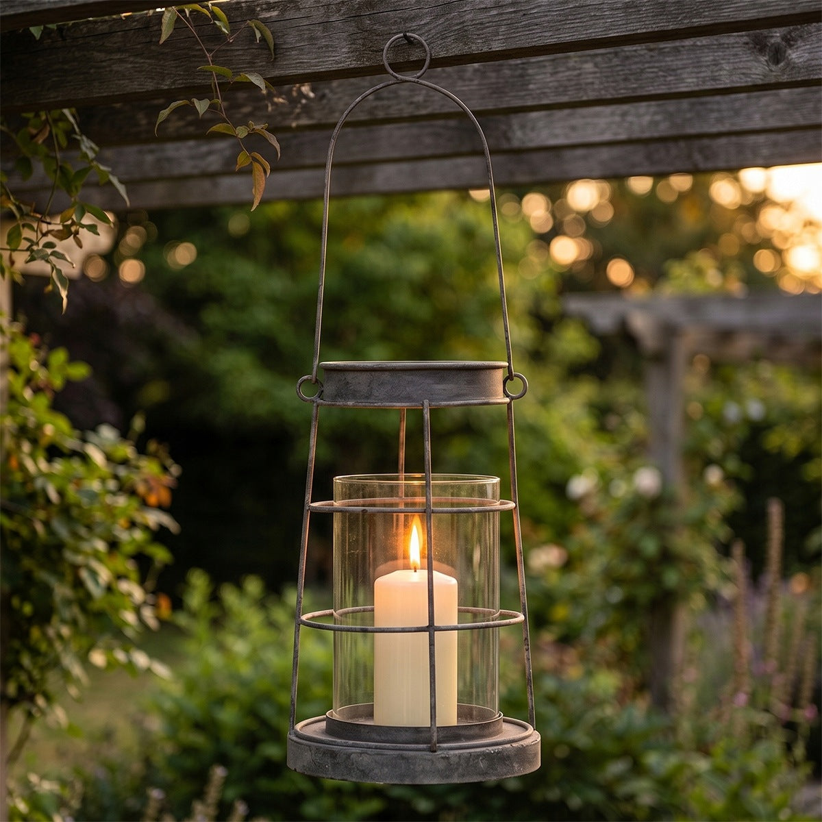 Klimatyczna aranżacja ogrodowa. Szary, metalowo-druciany lampion ze szklanym wkładem zawieszony jest na drewnianej belce pergoli za pomocą okrągłego ucha. Wewnątrz lampionu płonie jasna świeca blokowa. W tle widać malowniczo rozmyty ogród oświetlony ciepłymi promieniami zachodzącego słońca, co tworzy efekt świetlistych okręgów (bokeh) prześwitujących przez liście.