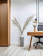 Nowoczesne, minimalistyczne biuro domowe (home office). Na jasnej podłodze, na tle białej ściany, stoi wysoki, metalowy wazon podłogowy w kolorze srebrnym, ozdobiony geometryczną fakturą. Wewnątrz wazonu znajduje się duży bukiet suchych gałęzi z drobnymi kwiatuszkami. Po lewej stronie widać fragment szafy z pionowymi, drewnianymi lamelami, a po prawej drewniane biurko, czarne krzesło biurowe oraz mały, beżowy wazonik.