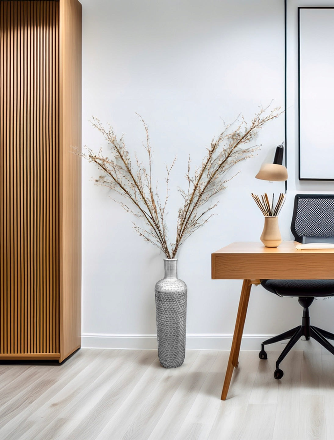 Nowoczesne, minimalistyczne biuro domowe (home office). Na jasnej podłodze, na tle białej ściany, stoi wysoki, metalowy wazon podłogowy w kolorze srebrnym, ozdobiony geometryczną fakturą. Wewnątrz wazonu znajduje się duży bukiet suchych gałęzi z drobnymi kwiatuszkami. Po lewej stronie widać fragment szafy z pionowymi, drewnianymi lamelami, a po prawej drewniane biurko, czarne krzesło biurowe oraz mały, beżowy wazonik.