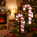 Zbliżenie na ubraną choinkę, na której wiszą dwie duże ozdoby w kształcie lasek cukrowych (candy canes) w brokatowe, czerwono-białe pasy. Laski owinięte są świecącym drucikiem LED, a obok nich luźno zwisają białe pudełeczka na baterie. W nastrojowym, rozmytym tle widać fragment salonu z zapalonym kominkiem i prezentami pod drzewkiem.