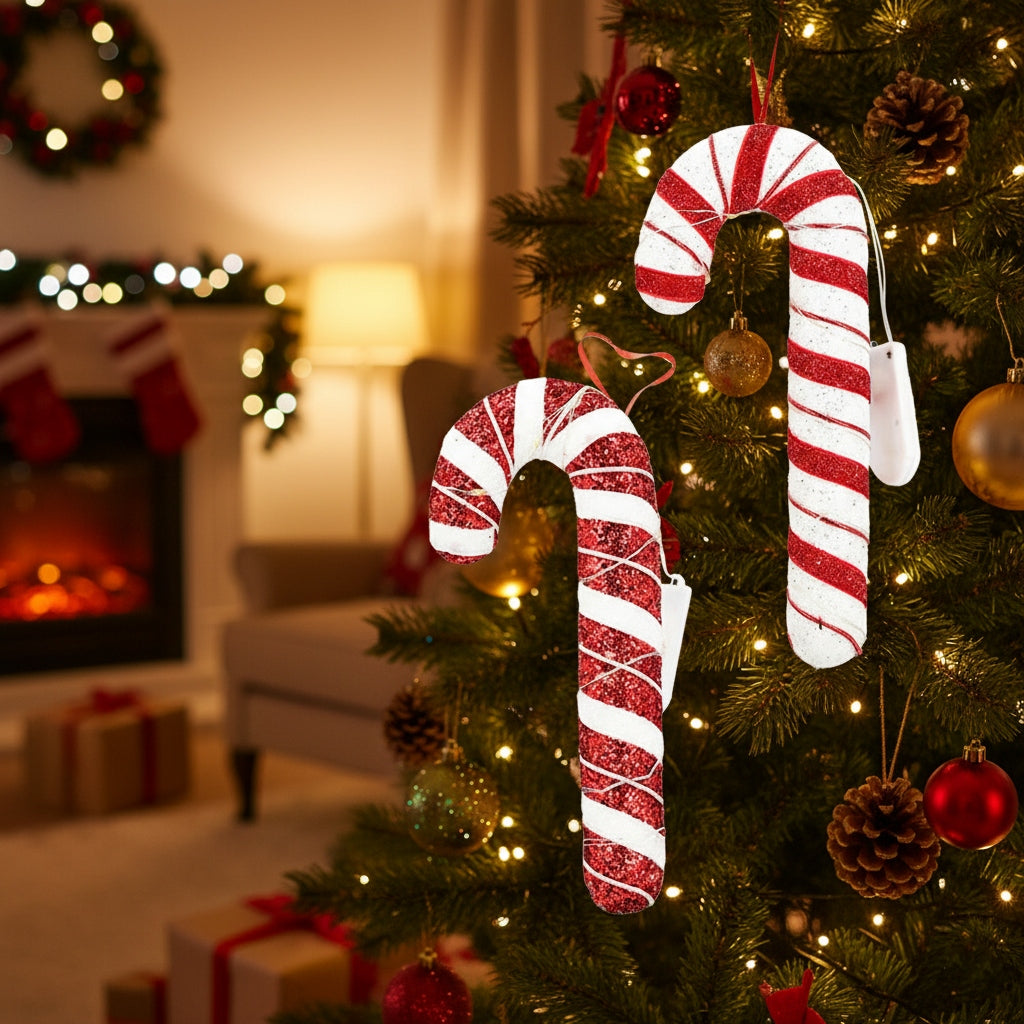 Zbliżenie na ubraną choinkę, na której wiszą dwie duże ozdoby w kształcie lasek cukrowych (candy canes) w brokatowe, czerwono-białe pasy. Laski owinięte są świecącym drucikiem LED, a obok nich luźno zwisają białe pudełeczka na baterie. W nastrojowym, rozmytym tle widać fragment salonu z zapalonym kominkiem i prezentami pod drzewkiem.