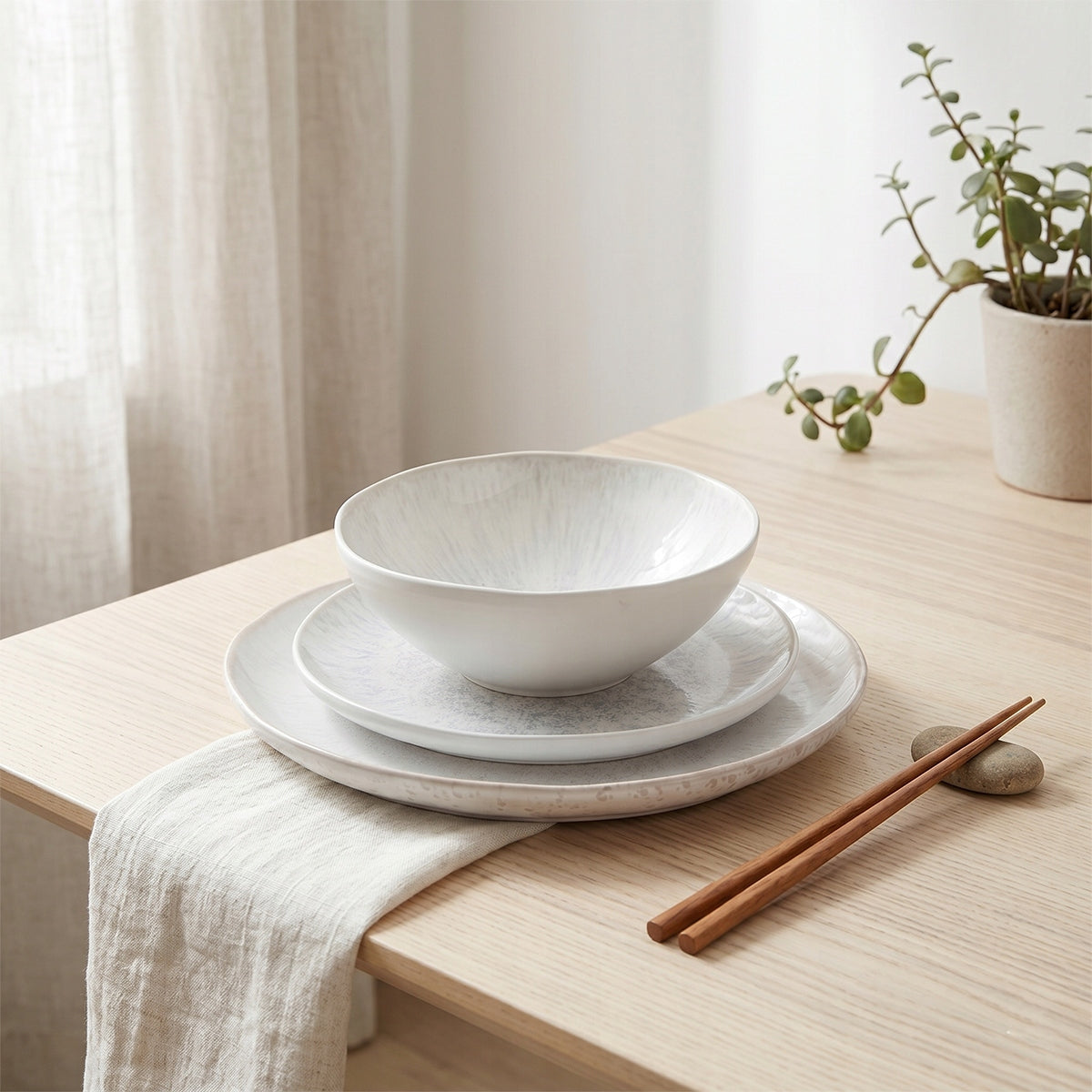 Spokojna, minimalistyczna kompozycja w stylu japandi. Na jasnym, dębowym blacie zaprezentowano trójelementowy zestaw ceramiczny: duży talerz obiadowy, mniejszy talerzyk oraz głęboką miskę. Naczynia mają piękny, jasny odcień z delikatnym, promienistym wzorem szkliwa. Aranżację dopełnia lniana serwetka, drewniane pałeczki oparte na drobnym kamieniu oraz zielona roślinka w surowej doniczce na dalszym planie, tworząc harmonijną, naturalną estetykę.