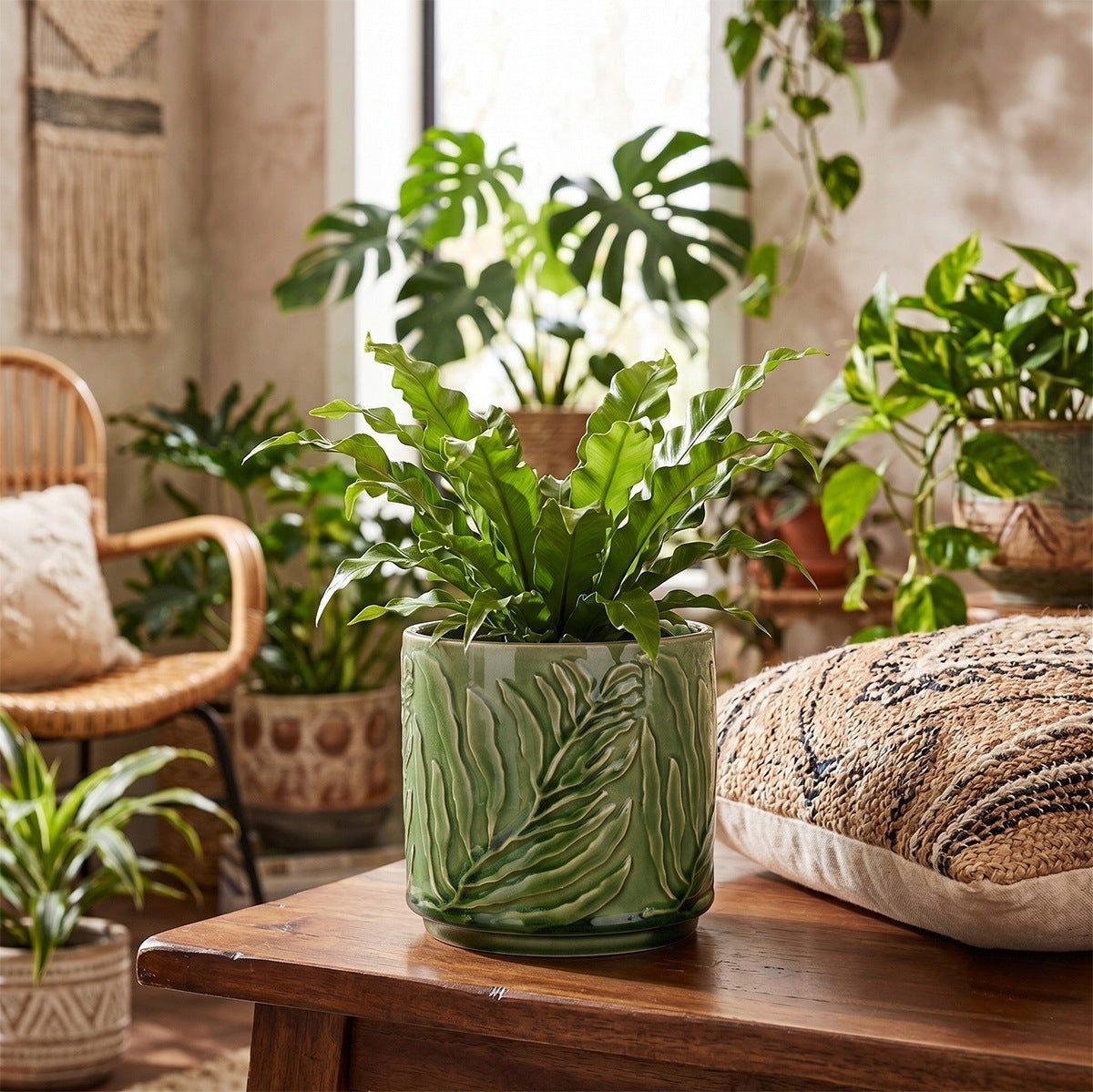 Roślinna aranżacja salonu w stylu urban jungle z elementami boho. Na drewnianym, rustykalnym stoliku kawowym stoi zielona, ceramiczna doniczka z wytłaczanym motywem liści, w której posadzono paproć o pofalowanych liściach (zanokcicę). Obok leży teksturowana poduszka w etniczne, plecione wzory. W tle pokoju znajduje się rattanowy fotel oraz bogata kolekcja innych roślin doniczkowych, w tym duża monstera dziurawa i gęste epipremnum.