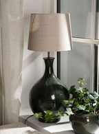 Ceramiczna lampa stołowa o pękatym, butelkowym kształcie, pokryta ciemnozielonym, błyszczącym szkliwem, zwieńczona jasnobeżowym, materiałowym abażurem. Lampa stoi na białym parapecie, skąpana w świetle dziennym. U jej podstawy wije się zielony bluszcz w ciemnej donicy, a po lewej stronie widać fragment jasnej, lnianej zasłony